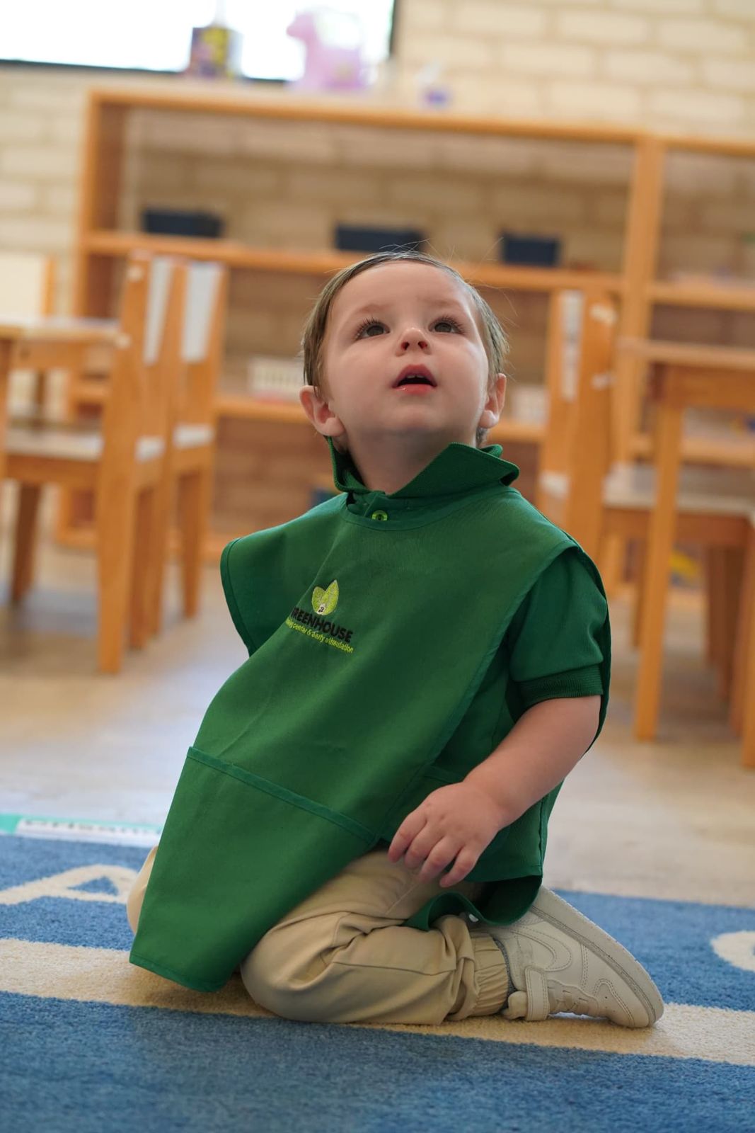 Niño de Pre-K con mandil verde de Greenhouse sentado en el tapete del salón