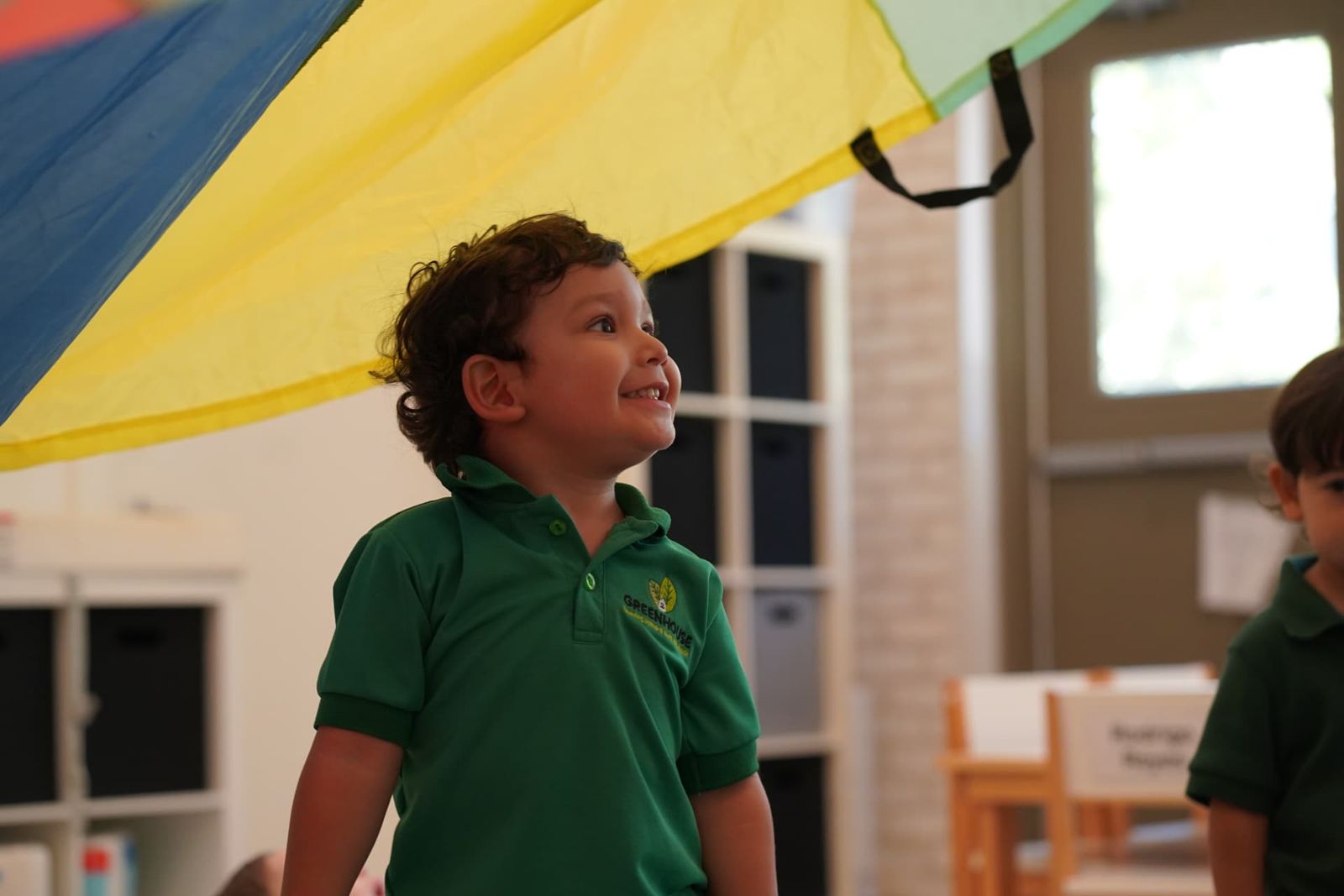 Niño sonriendo durante el juego del paracaídas en clase de preescolar en Greenhouse